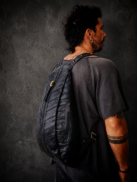 Male model wearing The Blackline Zip Pack against a dark textured wall, showing the black backpack, long front zipper, side pocket, and adjustable straps.