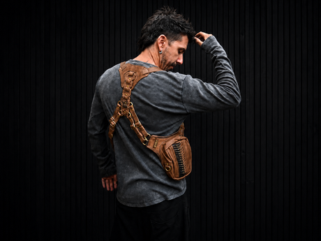 Male model wearing The Brown Prometheus leather shoulder harness bag from the back against a dark ribbed wall, showing the brown leather straps, brass hardware, and side pouch.