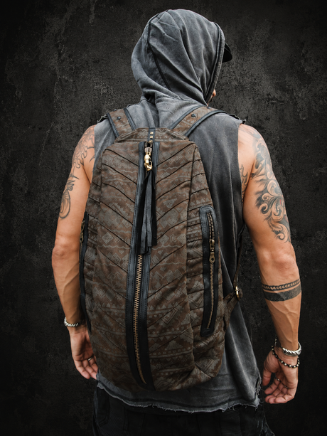 Male model wearing The Dustline Zip Pack against a dark textured wall, showing the brown grey geometric backpack, long front zipper, side pocket, and adjustable straps.