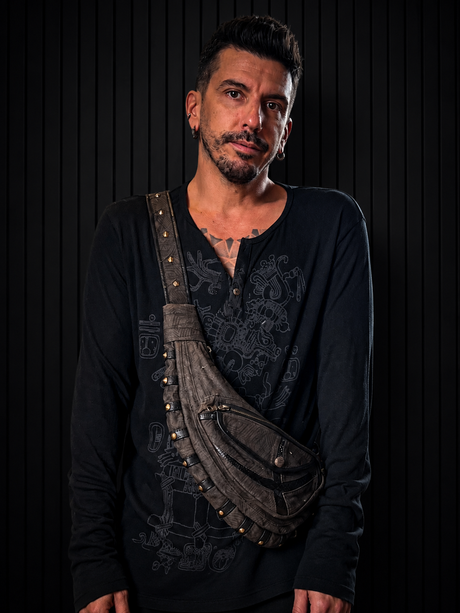 Male model wearing The Fabric Crescent Sling across the chest against a dark wood wall, showing the brown textured fabric body, black leather trim, brass studs, and curved sling shape.