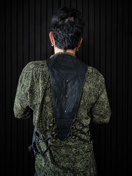 Back view of male model wearing a black leather shoulder harness bag against a dark vertical wall, showing the back panel, rivets, straps, and side pouch placement.