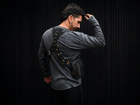 Male model wearing The Prometheus black leather shoulder harness bag from a side-back angle against a dark ribbed background.