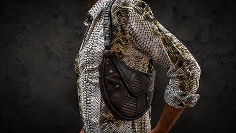 Male model wearing The Twin Orbit Bag as a dark brown leather crossbody utility pouch against a dark wall background
