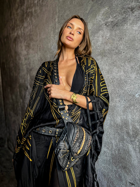 Female model wearing The Zenon waxed cotton hip pouch crossbody over a black outfit and black-and-gold draped layer against a textured gray wall.